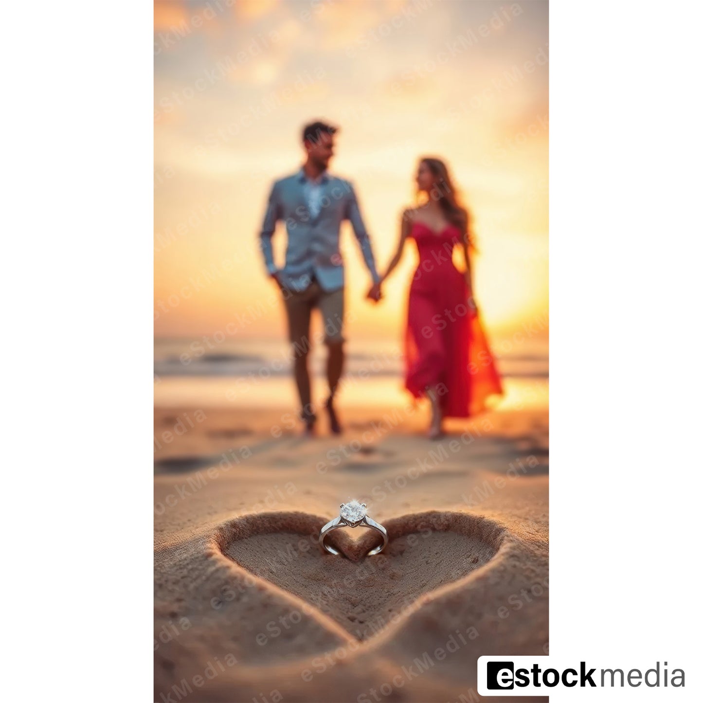 Diamond solitaire engagement ring resting inside heart drawn in beach sand with couple holding hands in soft focus background at golden sunset with man in light blue shirt and woman in flowing red off-shoulder dress