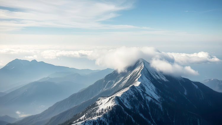 Aerial view of snow-capped alpine mountain peak with clouds and layered mountain ridges in blue haze