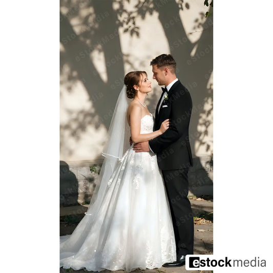 Romantic bride in white strapless wedding gown with flowing veil embracing groom in black tuxedo outdoors with artistic shadow patterns