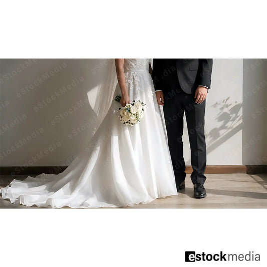 Elegant Caucasian bride in white strapless wedding gown with flowing train holding white flower bouquet standing beside groom in black suit indoors with dramatic shadows